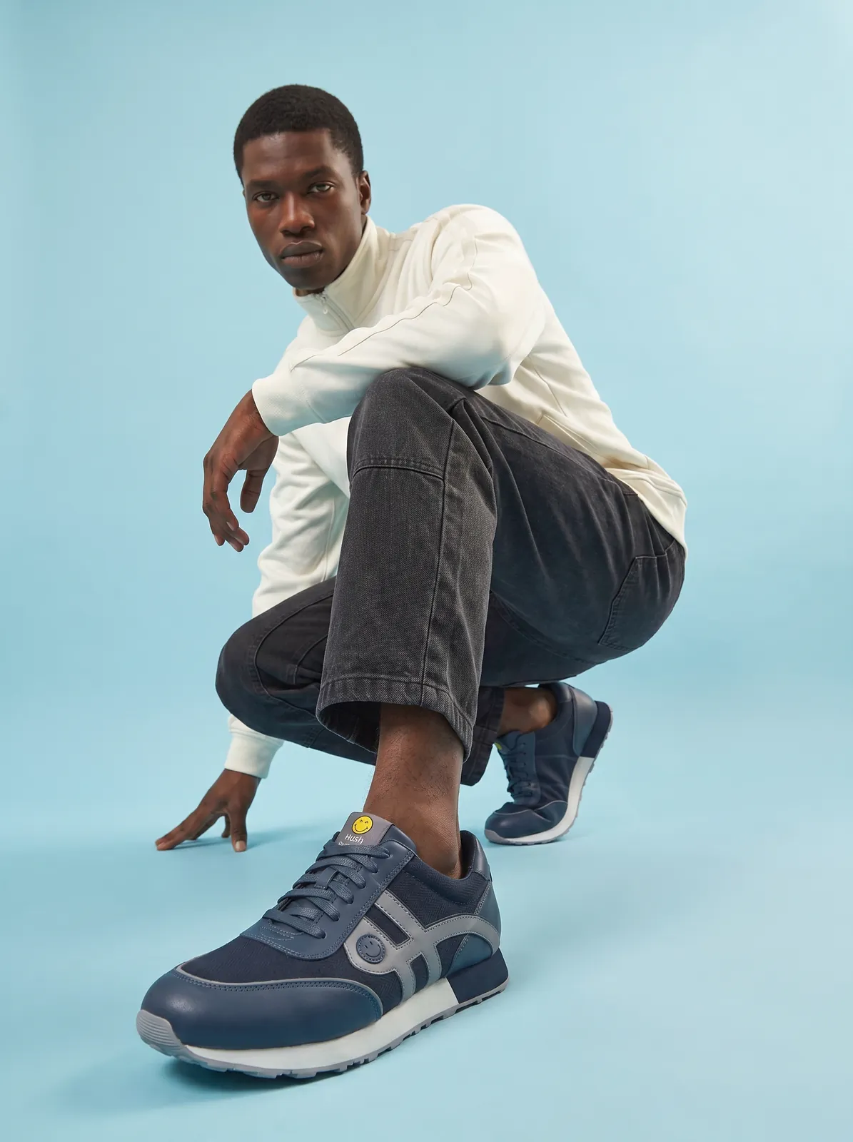 Male model crouching in navy running sneakers on a light blue background, footwear on-model photography